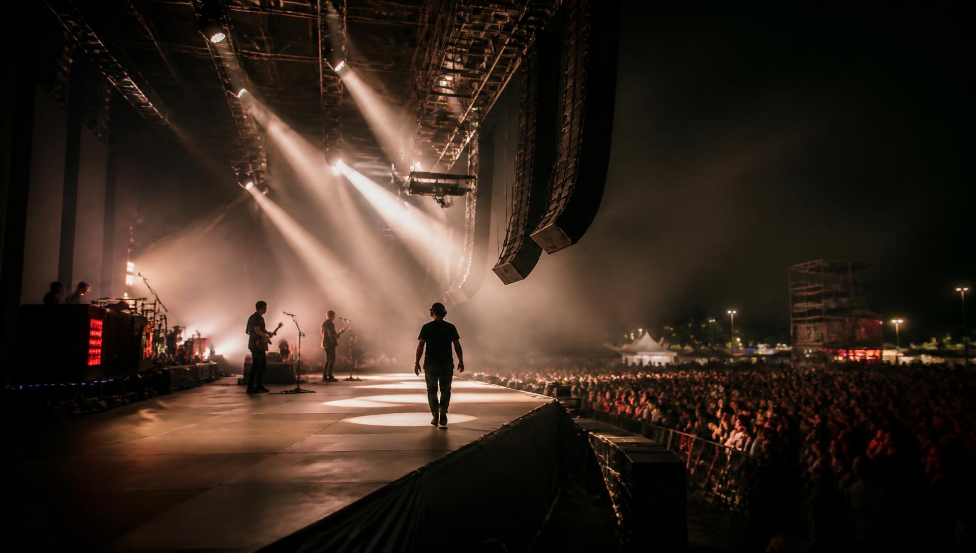 Crew member crossing a large outdoor event stage under lighting truss with a line array overhead and an audience below